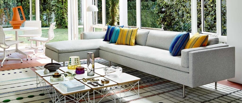 A modern living room scene featuring a light gray sectional sofa adorned with colorful striped cushions, a stylish coffee table with decorative objects, and large windows overlooking a green garden.