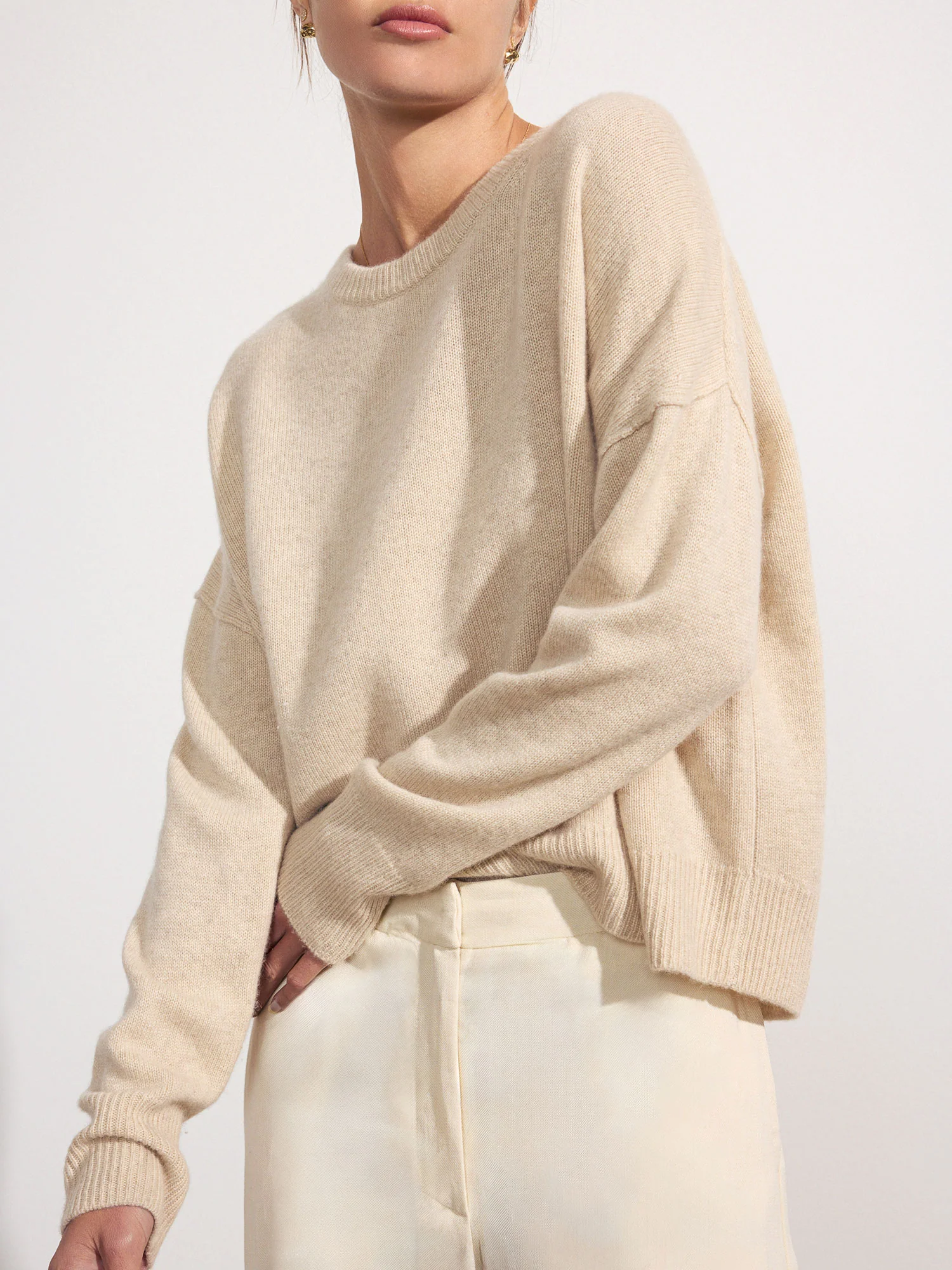 A close-up of a model wearing an oatmeal-colored cashmere sweater with a relaxed fit, paired with light linen pants. The sweater features a crew neck, slightly fitted sleeves, and a curved hem.