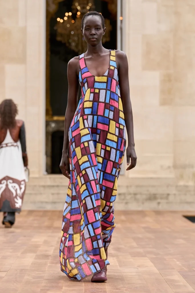 A model walks down the runway wearing a colorful patchwork dress with geometric patterns, showcasing vibrant hues of purple, yellow, red, and blue.