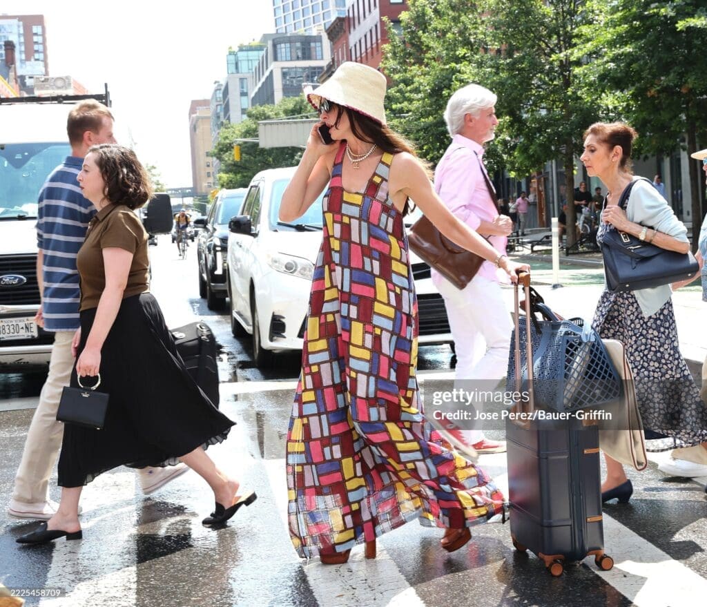 Actress Anne Hathaway in a Gabriela Hearst colorful patterned maxi dress carrying a suitcase and talking on her phone, walking through a busy street with other pedestrians.