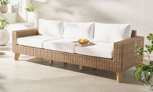 Contemporary outdoor sofa made of woven material with white cushions and a tray of oranges on it, surrounded by potted plants in a bright space.