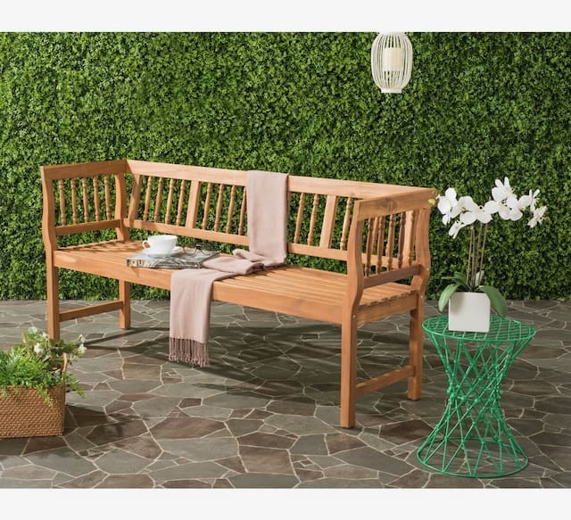 Outdoor wooden bench with a soft throw blanket draped over it, accompanied by a tea set and decorative table, surrounded by greenery.