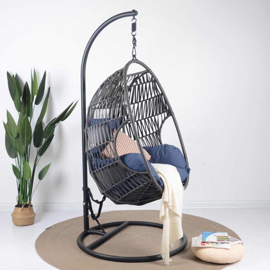 Modern hanging chair with a woven design and cushions, positioned beside a tall plant and a round mat, featuring a cozy throw and books on the floor.