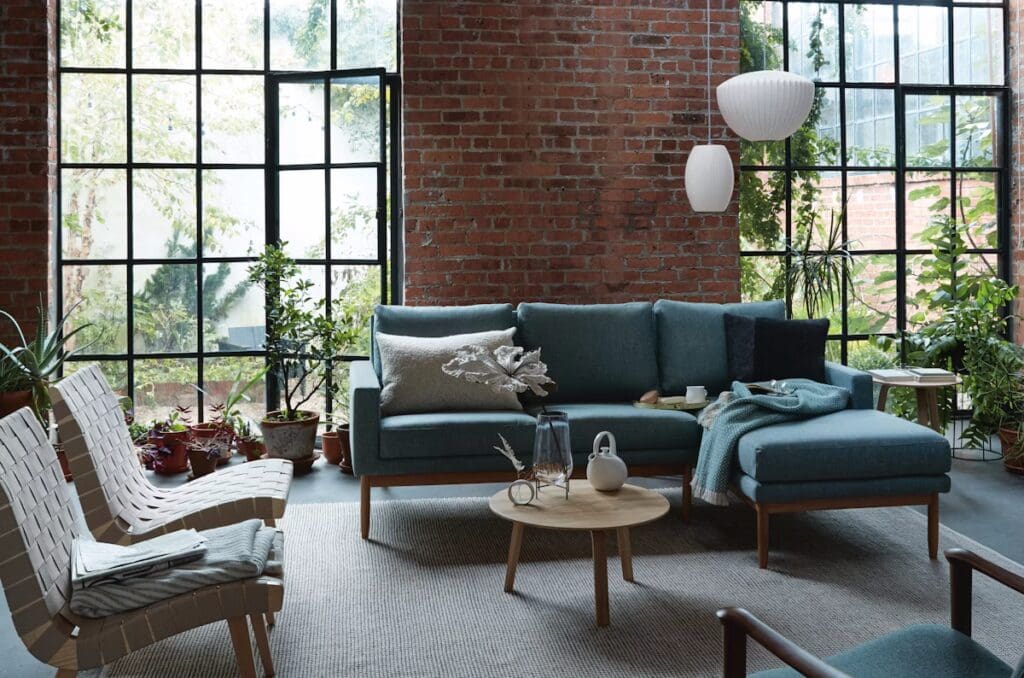 A modern living room with a blue sectional sofa, wooden coffee table, and woven chairs, surrounded by indoor plants and large windows with a view.
