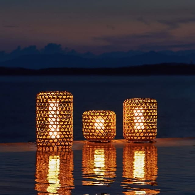 Three decorative lanterns with woven designs glowing softly by the water's edge during dusk, reflecting in the water with a serene ocean view in the background.