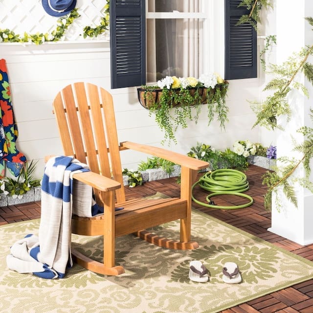 Wooden rocking chair placed on a decorative rug, accompanied by a blue and white blanket, flip-flops, and surrounded by potted plants and floral decorations.