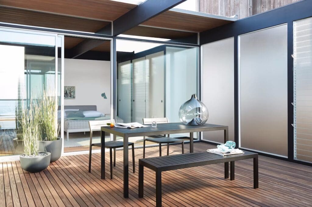 Modern outdoor dining area featuring a sleek black table and benches on a wooden deck, surrounded by minimalist decor and glass walls.