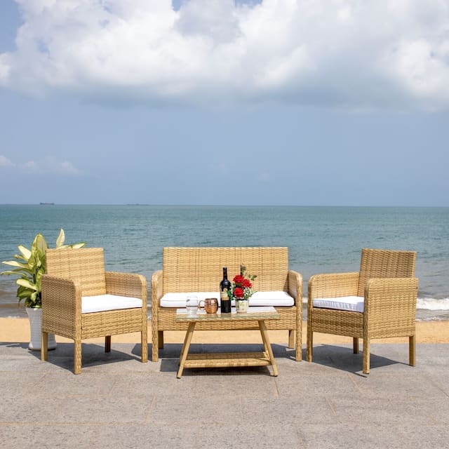 A cozy outdoor seating area featuring a wicker sofa and two armchairs, arranged around a small table with drinks and flowers, overlooking a serene ocean view under a partly cloudy sky.