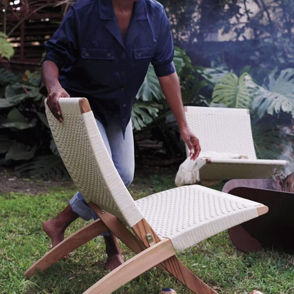 A person setting up a white woven camping chair in a green outdoor space surrounded by tropical plants.