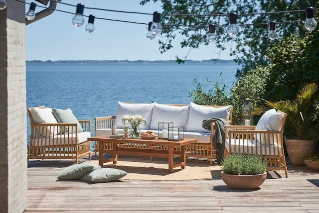 Stylish outdoor seating area featuring a wooden table and comfortable cushions, overlooking a serene body of water, adorned with potted plants and decorative lanterns.