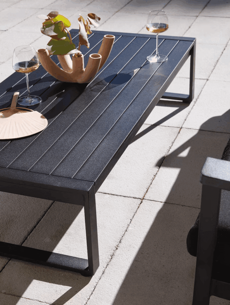 Close-up of a stylish outdoor coffee table made of dark material, featuring a unique vase with leaves and two wine glasses, set against pale tiled flooring.