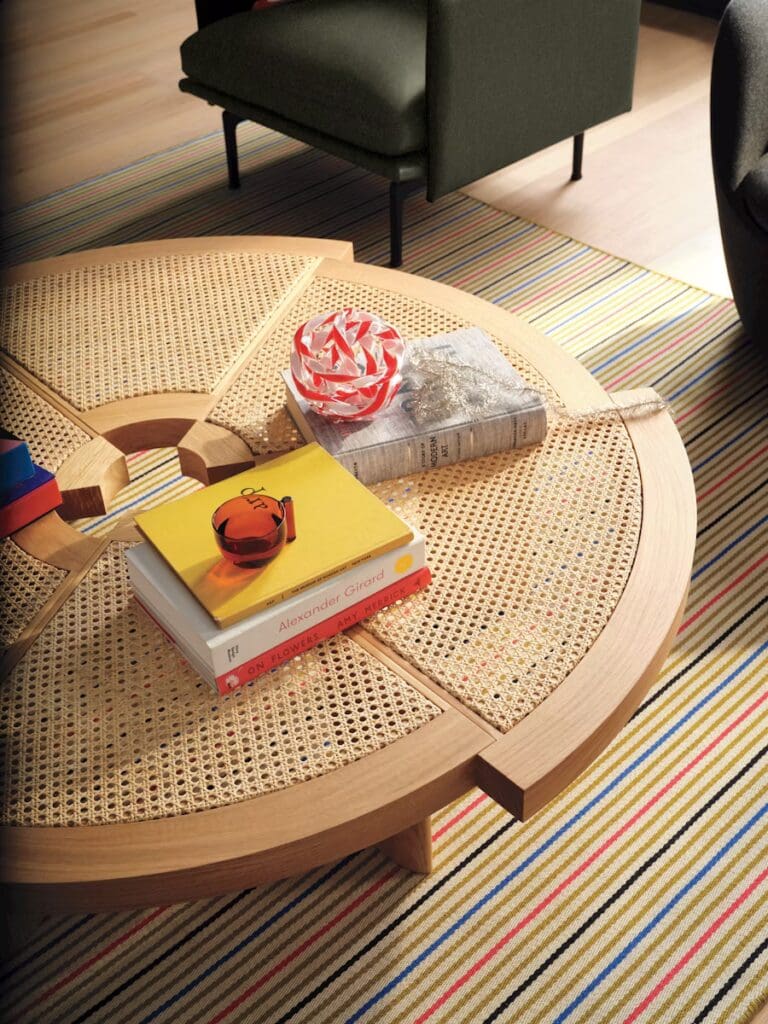 A modern low coffee table with a circular design made of wood and cane, displaying books and decorative items on top. In the background, a green armchair and a colorful striped rug can be seen.