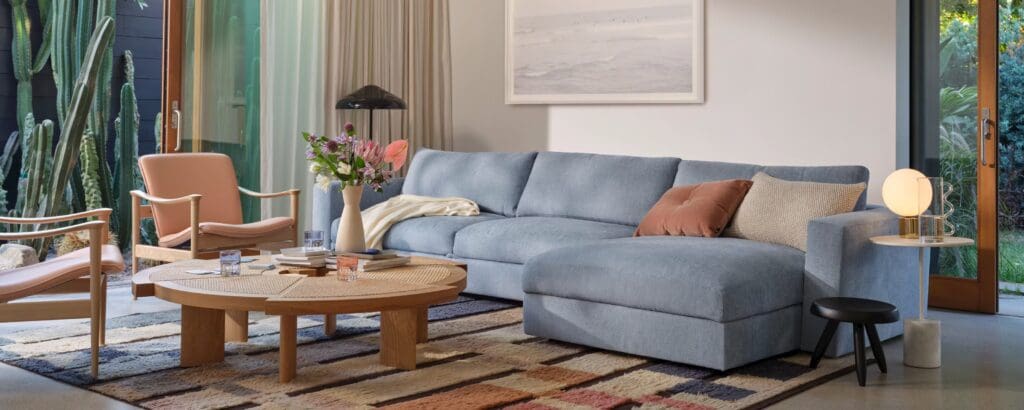 A modern living room featuring a blue sectional sofa with a chaise, a round wooden coffee table, stylish chairs, a floor lamp, and large windows showing plants outside.