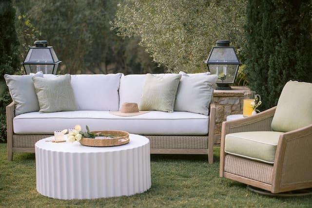 Stylish outdoor seating area featuring a comfortable sofa and armchair with green cushions, a round coffee table, and decorative lanterns. Lush greenery surrounds the space.