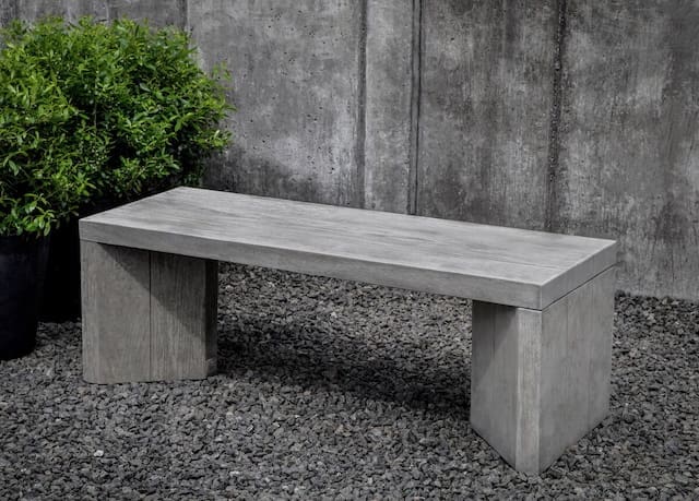 Modern concrete bench with clean lines, placed on a gravel surface next to a potted plant and a textured gray wall.