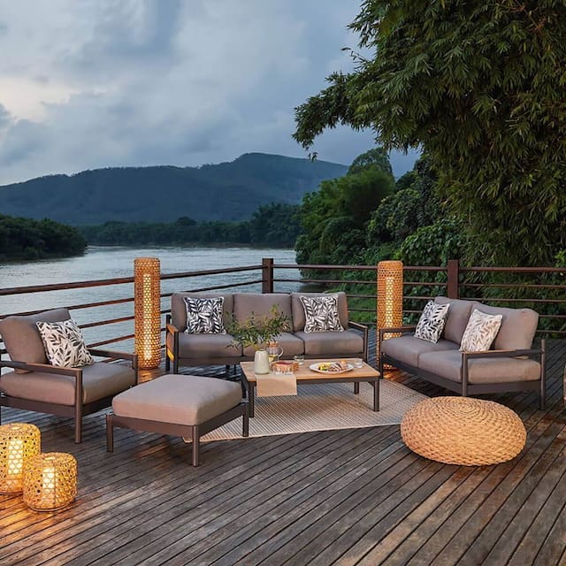 Cozy outdoor seating area with modern furniture overlooking a serene river, surrounded by lush greenery and mountains in the background. The space includes decorative lanterns, a coffee table with snacks, and a natural fiber rug.