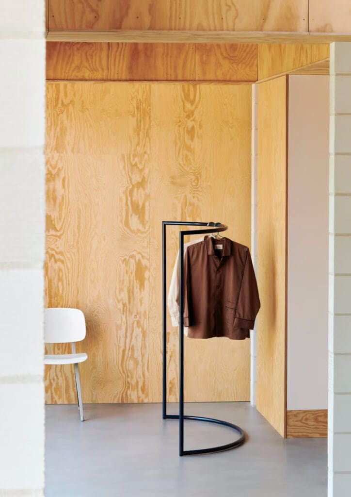 A modern interior featuring a wooden wall, a minimalist clothes rack with a brown jacket and a beige shirt hanging, and a white chair in the foreground.
