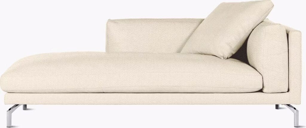 A modern chaise lounge in a light cream fabric with a minimalist design, featuring a single angled back cushion and a sleek metal base.