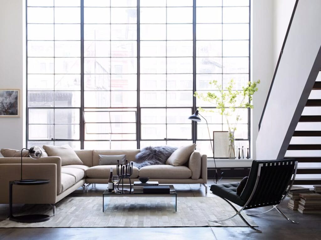 A modern living room with a beige sectional sofa, a glass coffee table, and a minimalist design. Features include a sleek floor lamp, decorative elements on a side table, and large windows allowing natural light, complementing the stylish decor.