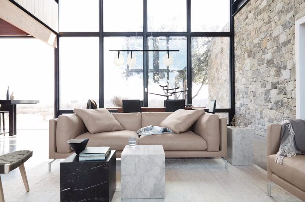 A modern living room with a light brown sofa, marble side tables, and large windows showcasing a natural view. The space features a minimalist design with stylish decor elements like a floor lamp and wooden accents.
