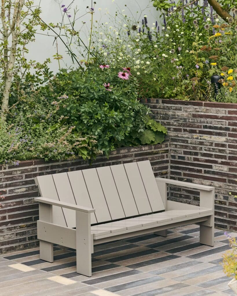 A modern outdoor bench made of light-colored wood sits on a patterned stone patio, surrounded by vibrant green plants and colorful flowers.