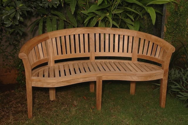Curved wooden outdoor bench made of teak, placed on grass surrounded by greenery.