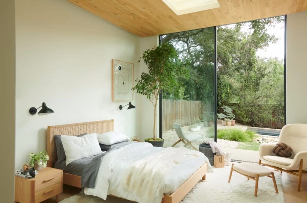 A cozy and modern bedroom featuring a wooden ceiling, a bed with a woven headboard, bedside table with a plant, and wall-mounted lamps. Large windows provide a view of a lush outdoor garden.