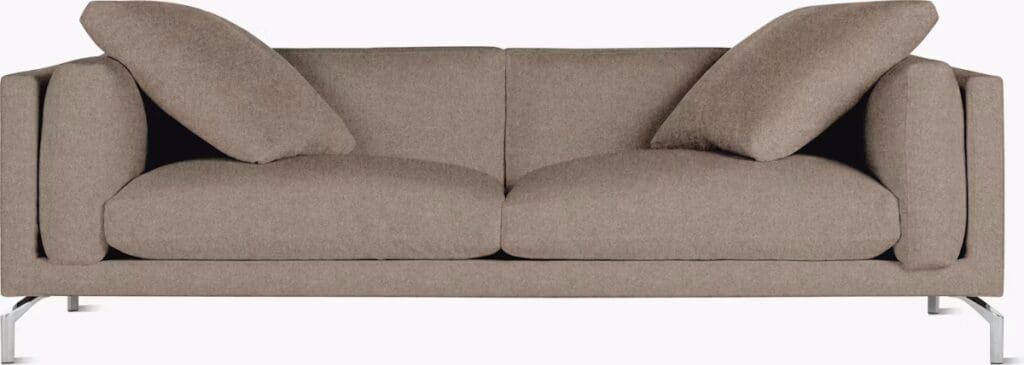 A modern, minimalist sofa with two cushions partially leaning against the backrest, showcasing a neutral brown fabric and sleek metal legs.