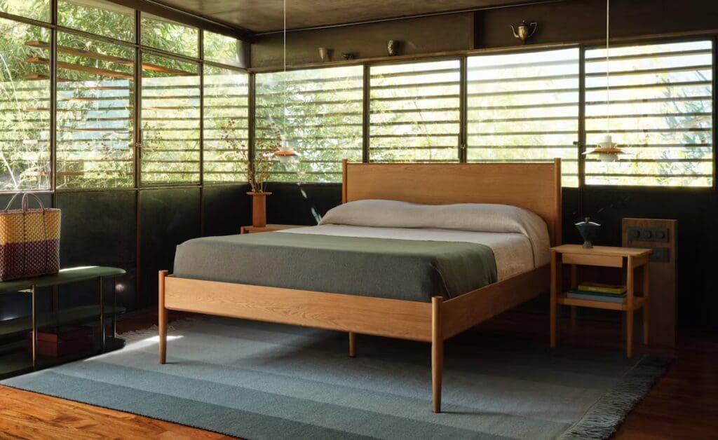Cozy bedroom featuring a wooden bed frame with a green bedspread, a matching nightstand, and large windows with horizontal slats allowing natural light to enter.