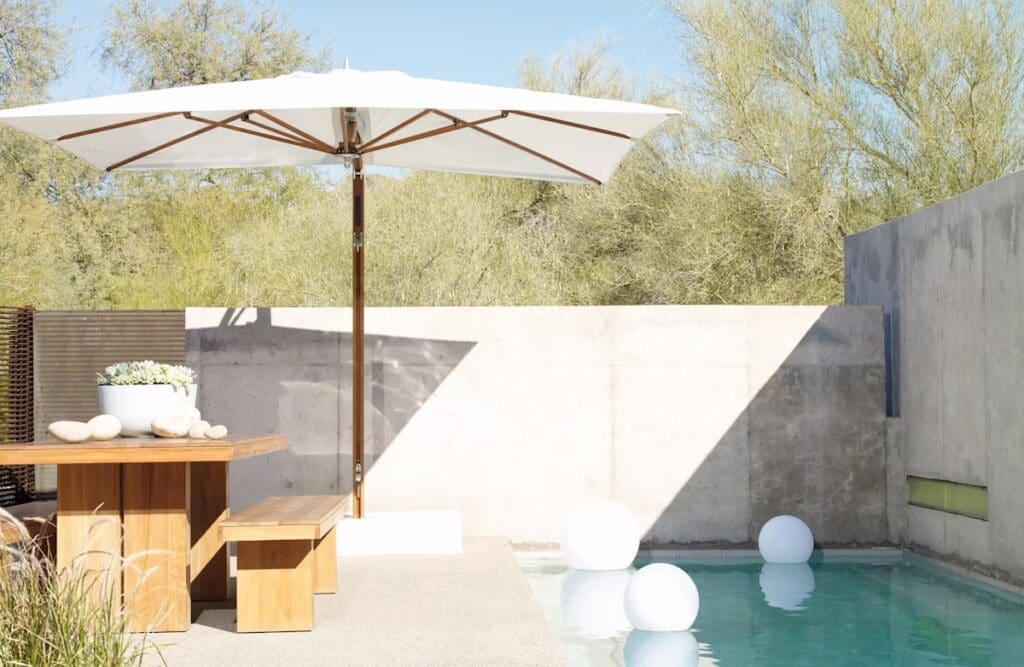 A modern outdoor space featuring a wooden table and benches beneath a large white umbrella, with a swimming pool and decorative round floats in the water, surrounded by greenery.
