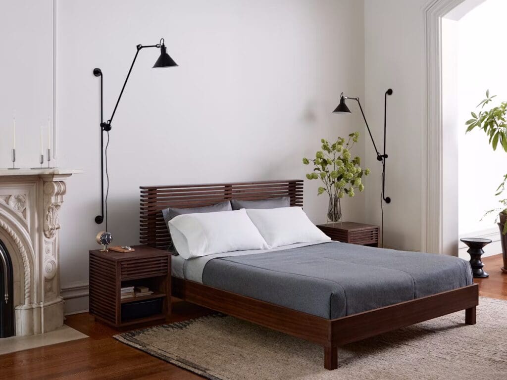 A stylish bedroom featuring a wooden bed frame, a gray blanket, and white pillows. There are modern bedside lamps and a decorative plant beside the bed, with an elegant fireplace on one side.