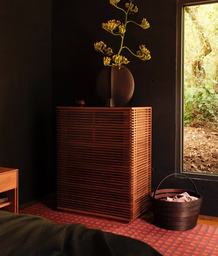 A stylish wooden cabinet with horizontal slats, topped with a round vase holding vibrant flowers, set against a dark wall and a window showcasing a natural landscape.