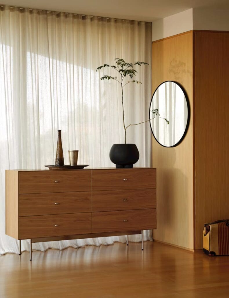 A stylish modern living room corner featuring a wooden chest of drawers with metallic handles, a sleek black vase with a plant, and a round mirror on the wall, illuminated by soft natural light filtering through sheer curtains.