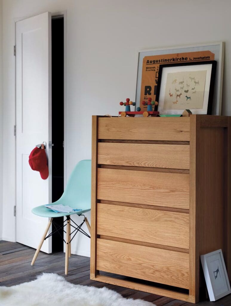 A minimalist bedroom interior featuring a wooden dresser with multiple drawers, a light blue chair beside it, and a door leading to another room. Decor items include a framed illustration on top of the dresser and a red hat hanging on the door.