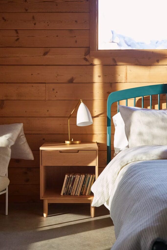 A cozy bedroom scene featuring a wooden wall, a teal bed frame with white bedding, a bedside table with books and a modern lamp.