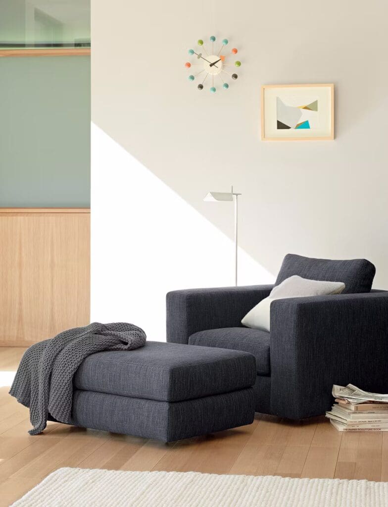 A modern living room setup featuring a dark upholstered armchair with a decorative pillow, a matching ottoman draped with a cozy blanket, a minimalist wall clock with colorful accents, and a framed artwork above the chair.