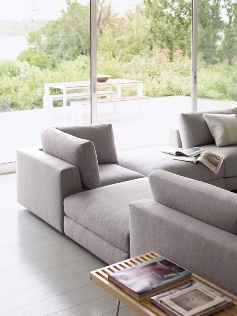 A contemporary living room featuring a light gray sectional sofa next to large windows with a view of greenery outside. A wooden side table with magazines is visible.