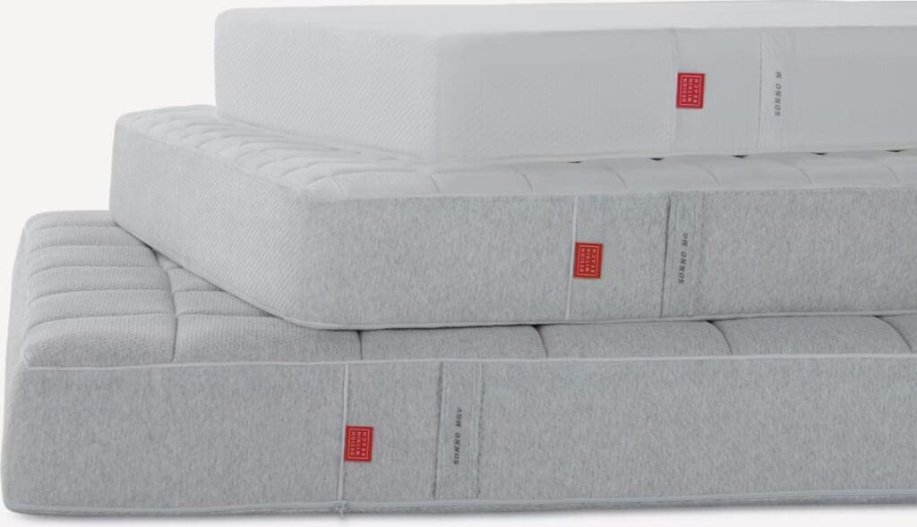 Three stacked mattresses in varying thicknesses, with a minimalist design and grey fabric covers, displayed against a white background.