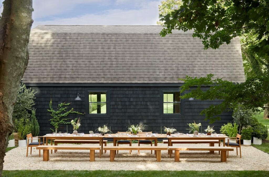Outdoor dining setup with a long wooden table and benches, surrounded by greenery and a black shingled house in the background.