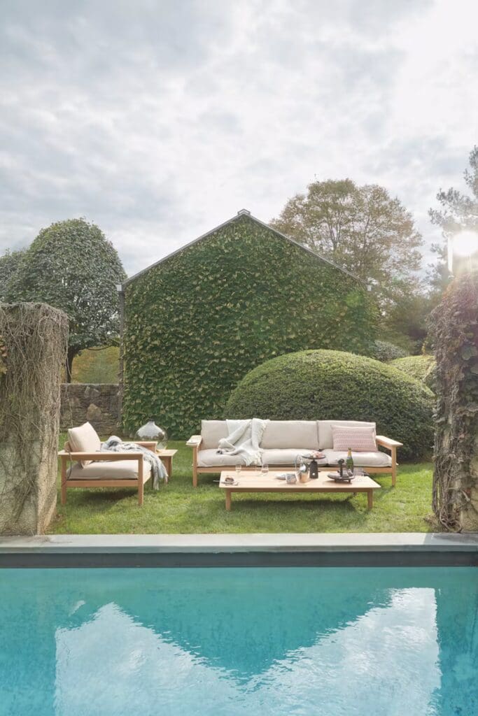 A serene outdoor living space featuring a comfortable sofa and wooden coffee table next to a pool, surrounded by lush greenery and a background of an ivy-covered house.