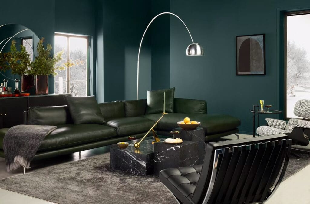 A modern living room featuring a dark green wall, a sleek green sectional sofa, a round floor lamp, and a black marble coffee table with decorative items. A large window offers a view of a snowy landscape.