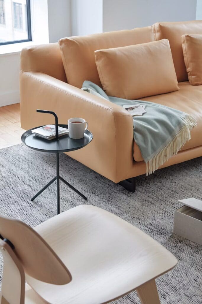 A cozy modern living room with a light-colored leather sofa, decorative pillows, and a soft throw blanket. A small black side table holds a coffee cup and magazines. A wooden chair is partially visible in the foreground, complementing the stylish decor.