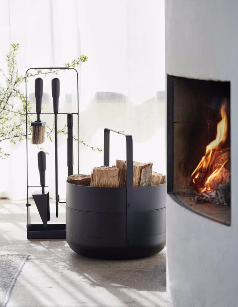 A modern firewood holder filled with logs, positioned next to a fireplace. Behind it, a set of fireplace tools stands on a concrete floor, with a bright, airy atmosphere from natural light filtering through white curtains.