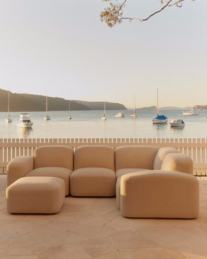 A modern, beige sectional sofa positioned outdoors with a serene waterfront view featuring sailboats in the background.
