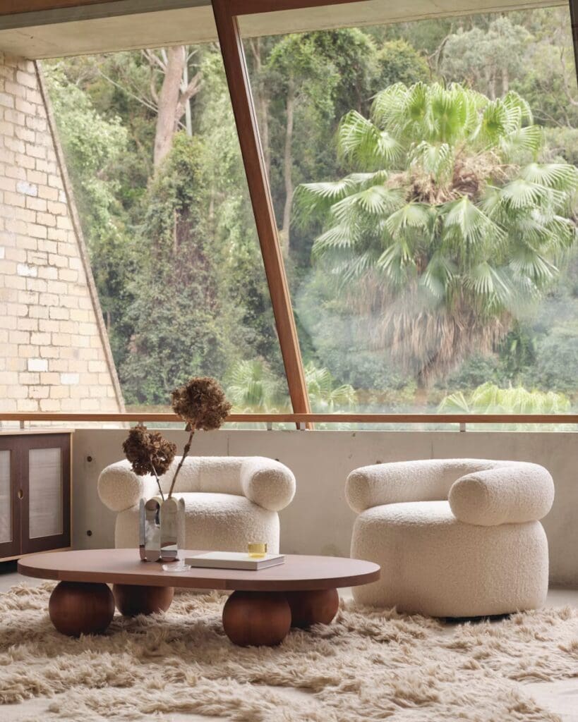 A cozy living room featuring two plush, round armchairs with a fluffy texture, a wooden coffee table with a unique base, and a decorative vase with dried flowers. The background includes large windows showcasing lush greenery.