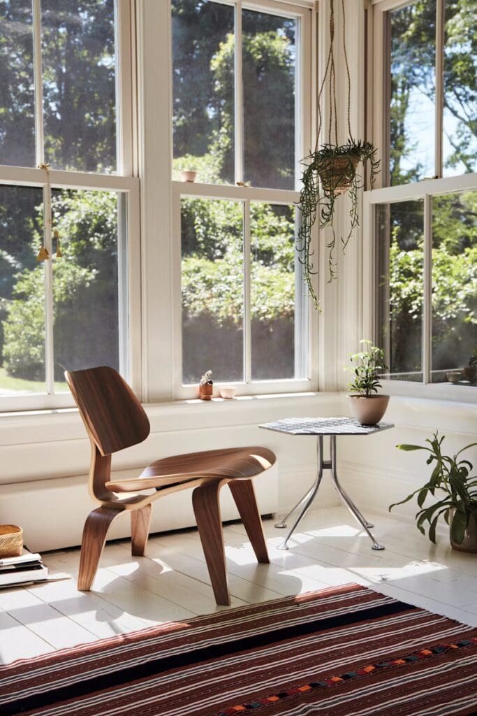 A stylish corner of a modern living room featuring a wooden chair, a small round table, large windows letting in natural light, and decorative plants. A patterned rug adds warmth to the space.