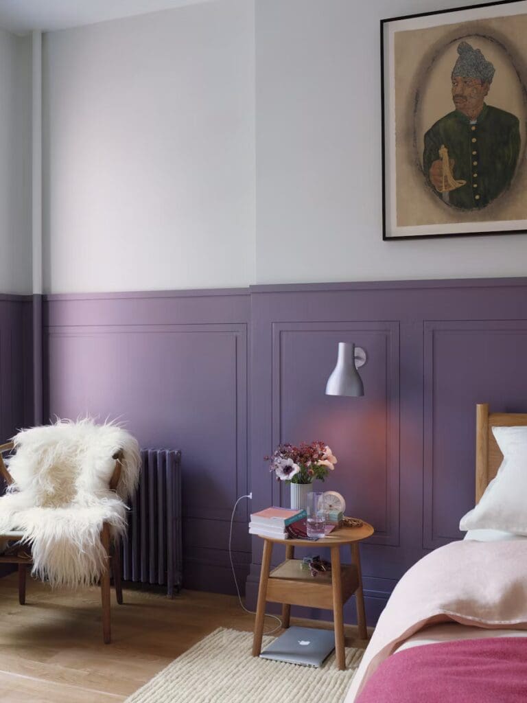 A cozy bedroom with a purple accent wall, featuring a wooden bed, bedside table with books and flowers, a plush white chair, and a framed portrait on the wall.
