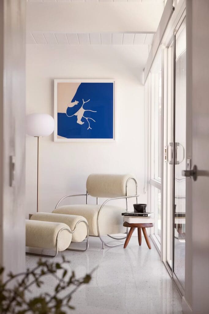 A modern living space featuring a curved lounge chair and ottoman in light fabric, accompanied by a small wooden side table holding a cup and book. A large abstract blue artwork is displayed on the wall, and large glass doors allow natural light into the room.