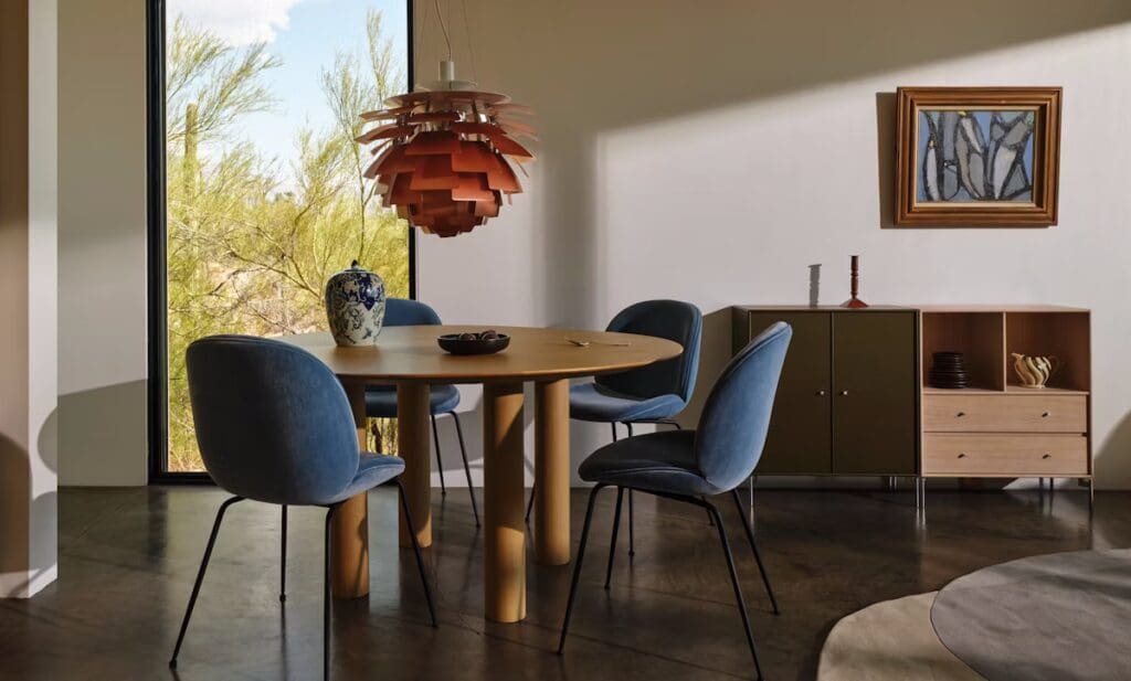 A modern dining area featuring a round wooden table surrounded by blue upholstered chairs. A stylish pendant light hangs above the table, and a decorative vase is placed in the center. In the background, large windows offer a view of greenery outside, complemented by a cabinet with drawers and a framed artwork on the wall.
