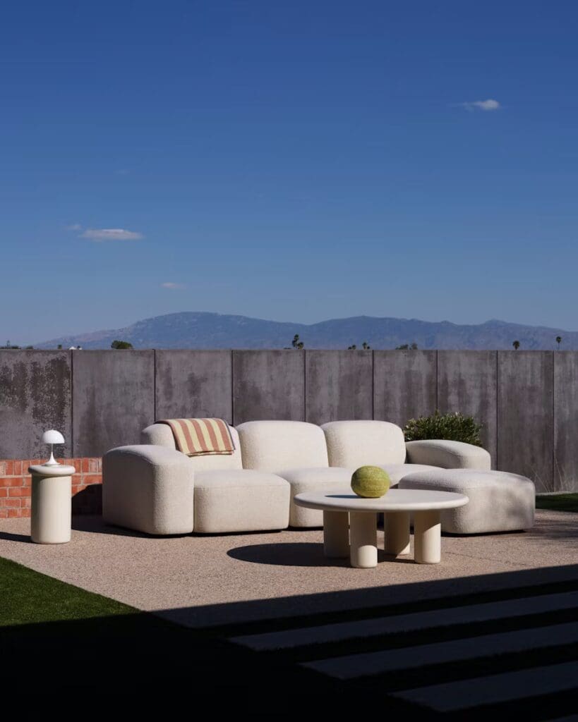 A modern outdoor seating arrangement featuring a plush cream-colored sofa with a striped pillow, paired with a round coffee table, against a backdrop of a concrete wall and clear blue sky.